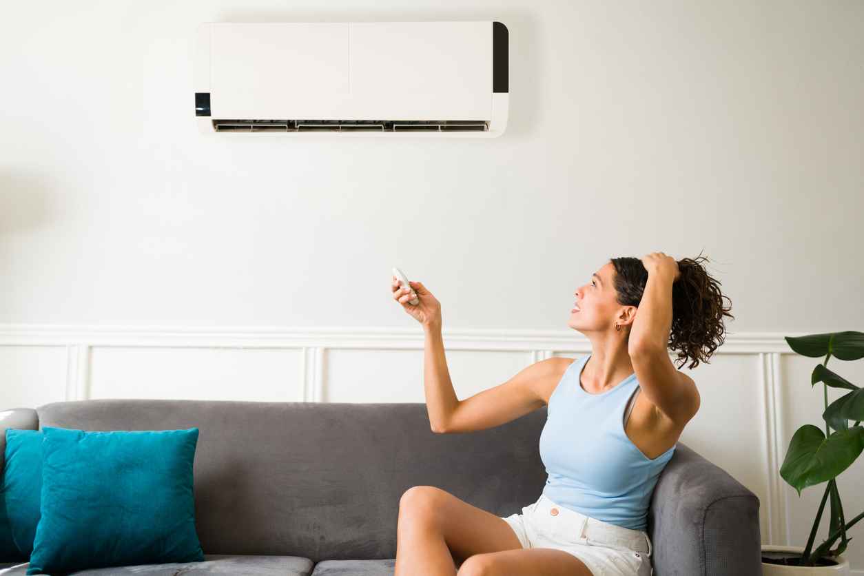 A woman sitting on her sofa at home sweating because her AC is not working. She is holding her hair up and trying to turn on her ductless mini split.
